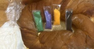 Undecorated king cake with sealed icing pouch and purple, green and gold decorating sugars.